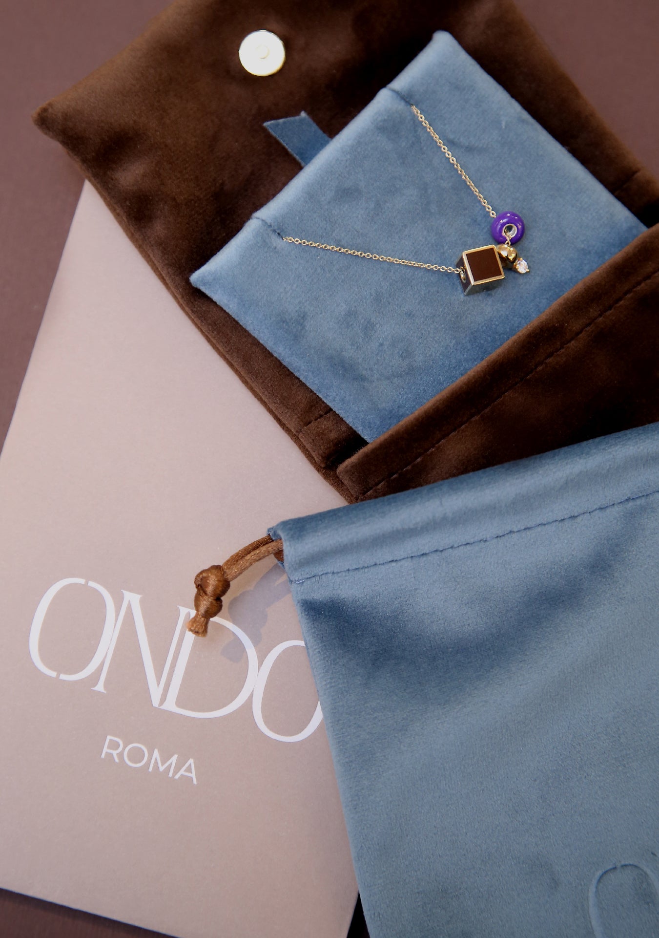 ONDO necklace with cube and charm details, elegantly placed on a blue velvet pouch inside brown packaging, with ONDO ROMA branding—showcasing refined presentation.