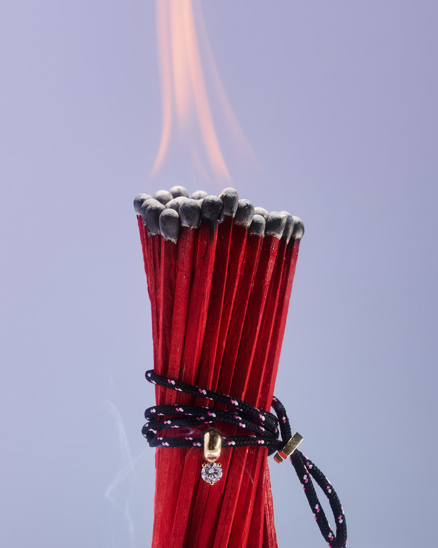 ONDO black cord bracelet with gold and diamond detail wrapped around a bundle of lit red matches—bold contrast showcasing the brand’s edgy, refined design.
